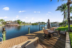A wooden deck with outdoor furniture overlooking a calm river, surrounded by greenery and a clear sky.