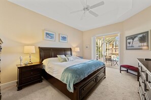 A bedroom with a bed, nightstands, a dresser, and a ceiling fan. There are framed pictures on the wall and a view of the outside through a door.