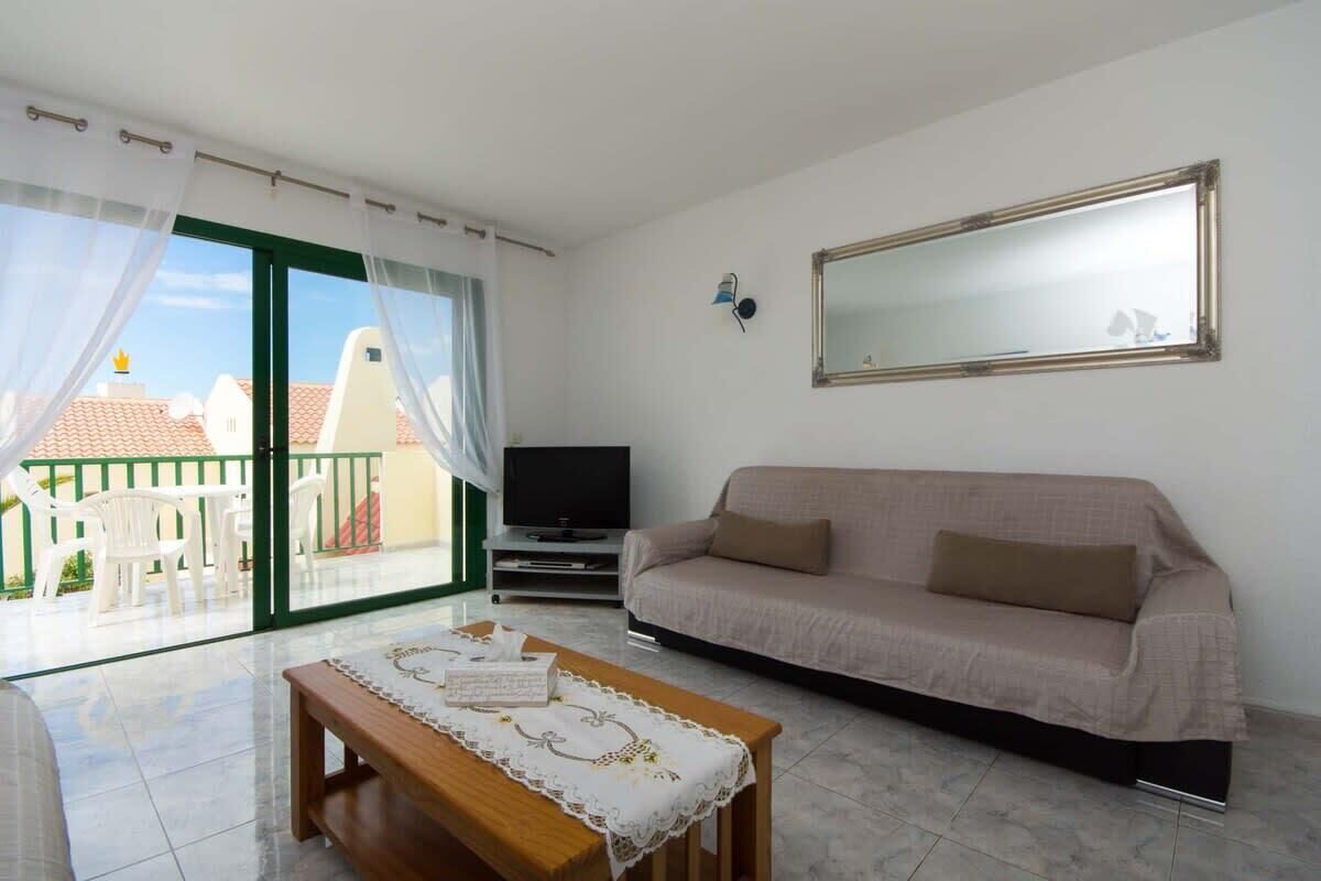 Un salon comprenant un canapé, un téléviseur, une table en bois recouverte d’une nappe et un balcon avec des chaises blanches.