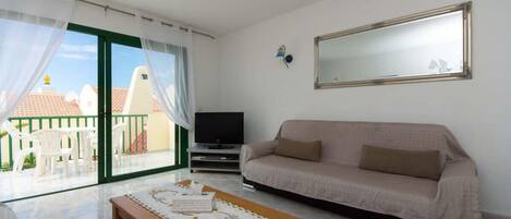 Un salon comprenant un canapé, un téléviseur, une table en bois recouverte d’une nappe et un balcon avec des chaises blanches.