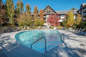 Une piscine dotée d’une terrasse en bois, entourée d’arbres et bordée d’un bâtiment dont le toit a la forme d’un triangle.