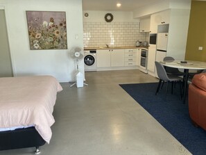 A compact room with a bed, a dining table with chairs, a kitchen area with a washing machine, and a small fan.