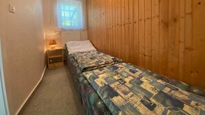 Quarto pequeno com uma cama de solteiro, parede de madeira, janela com cortinas finas, mesa de cabeceira com abajur e piso coberto por carpete.