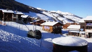 A snow-covered mountain village with wooden buildings, a ski lift, and a clear blue sky.