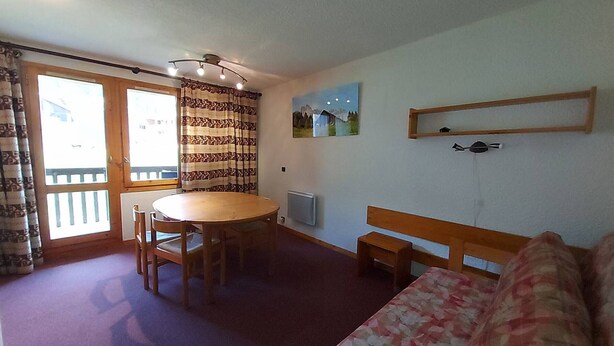 A room with a dining table, chairs, a sofa, a window with curtains, a radiator, and a wall-mounted shelf.