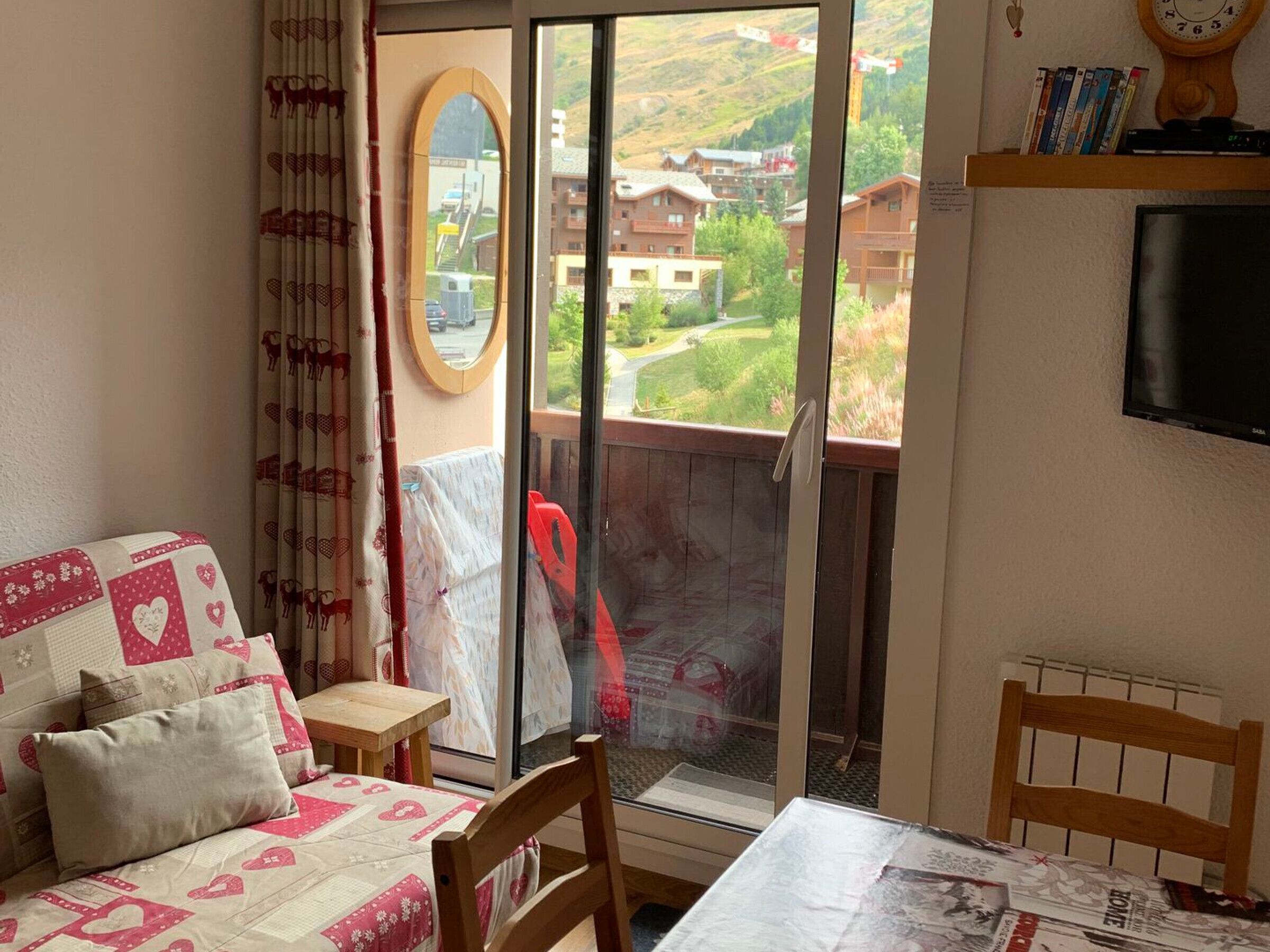 Ein gemütliches Zimmer mit Bett, kleinem Tisch, Stuhl, Fernseher und einem Balkon mit Blick auf Gebäude und Grünflächen.