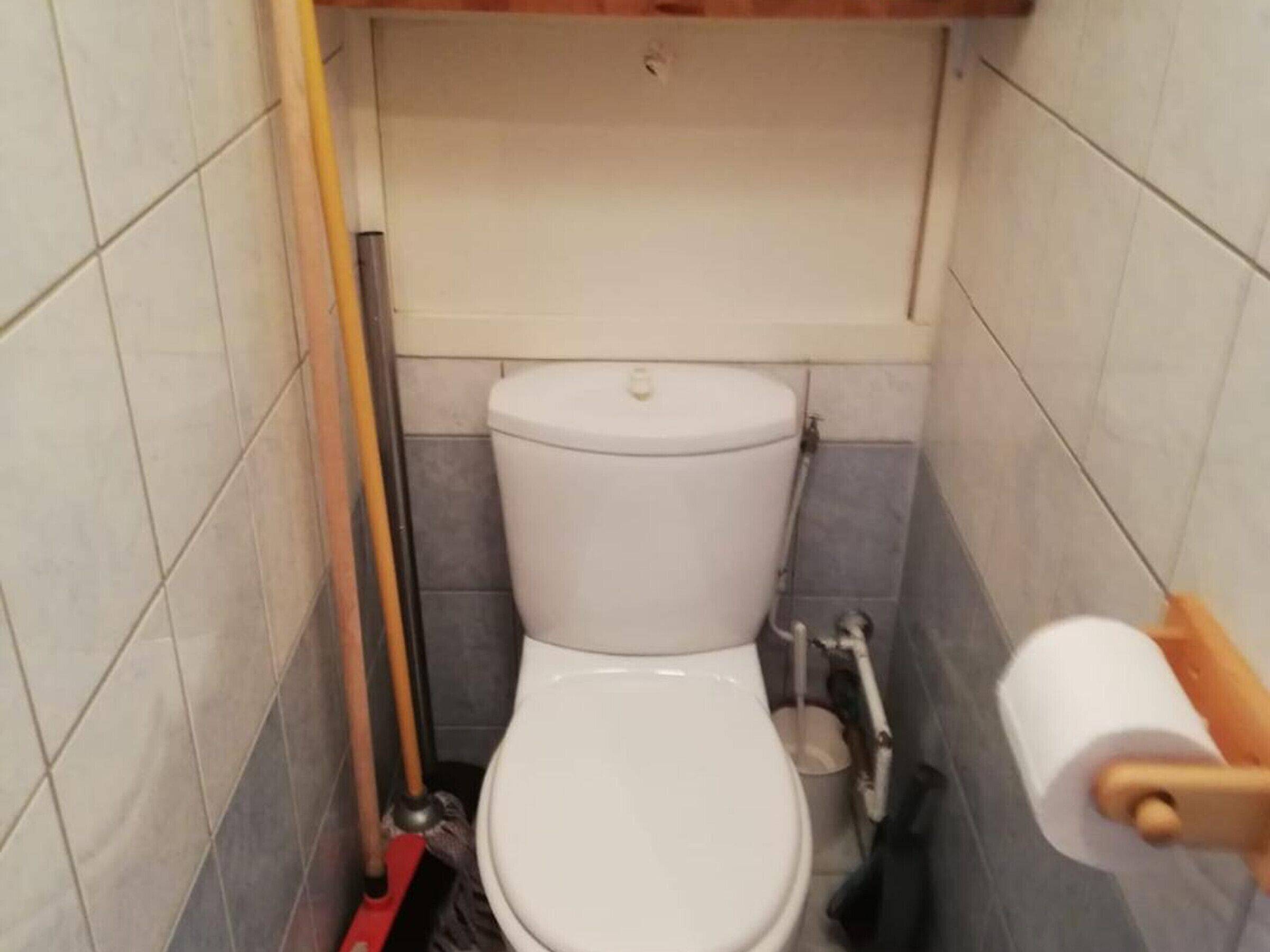 Une petite salle de bain carrelée, équipée de toilettes, d’un balai et d’un porte-rouleau de papier toilette.