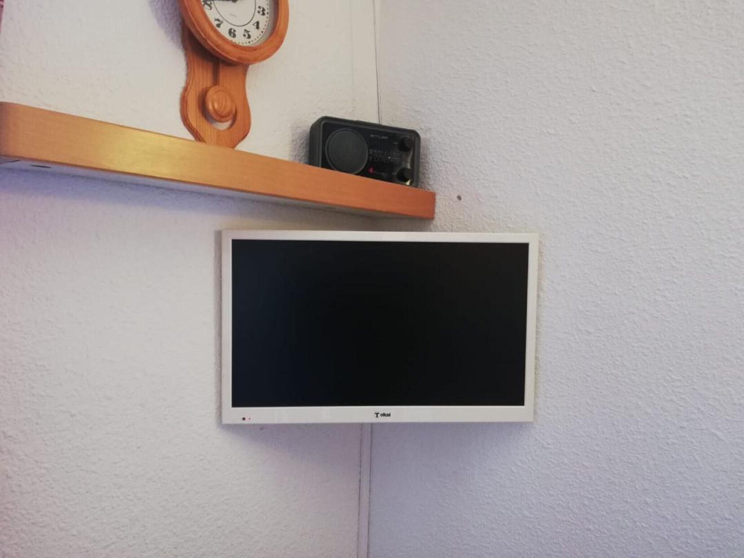 Un téléviseur blanc fixé au mur, une étagère en bois avec une horloge au-dessus, et un petit appareil noir posé sur l’étagère.