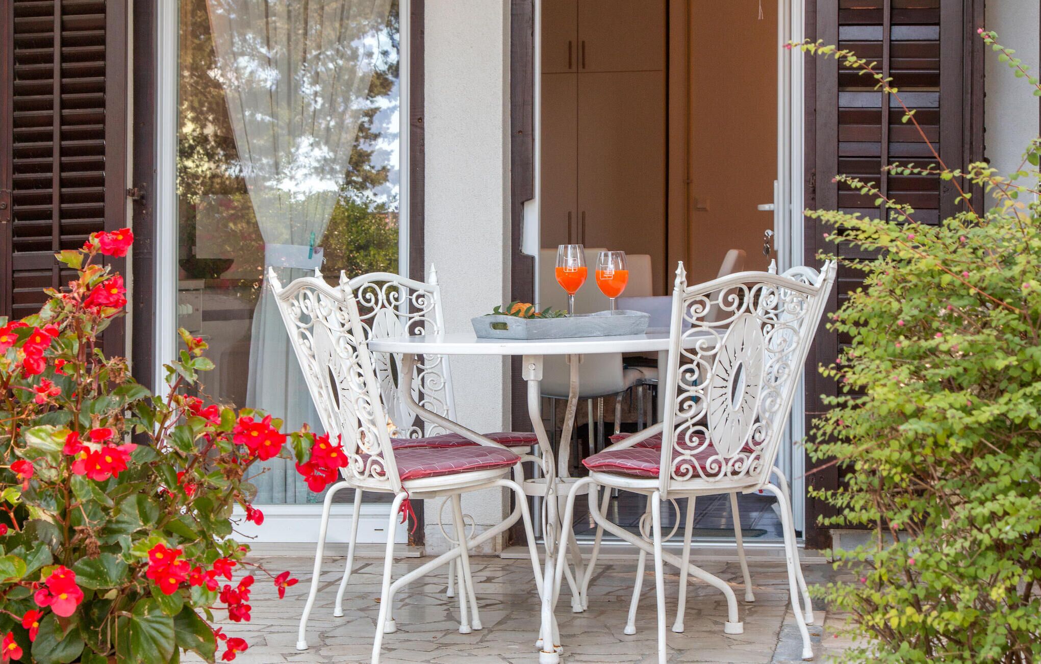 Een terras met een ronde tafel en vier witte stoelen met rode kussens. Op de tafel staan twee glazen oranje drank.