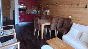 L’intérieur d’un chalet accueillant, avec un plafond en bois, un coin repas comprenant une table et des chaises en bois, une cuisine aux placards rouges et un canapé blanc.