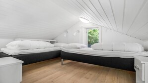 Une petite chambre mansardée, aux murs blancs, avec deux lits, une fenêtre et un plancher en bois.