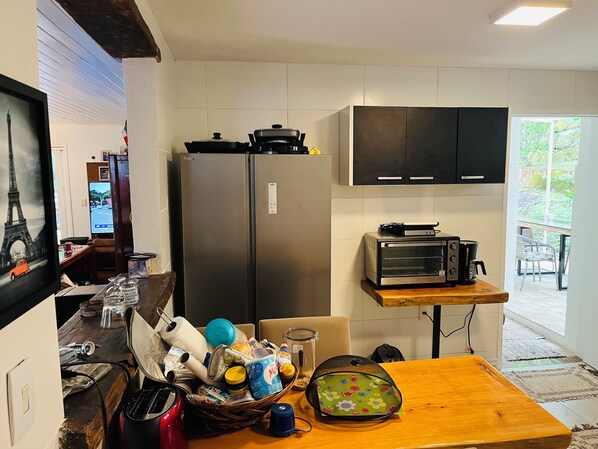Una cocina compacta con refrigerador, horno tostador y una mesa de madera llena de objetos varios.