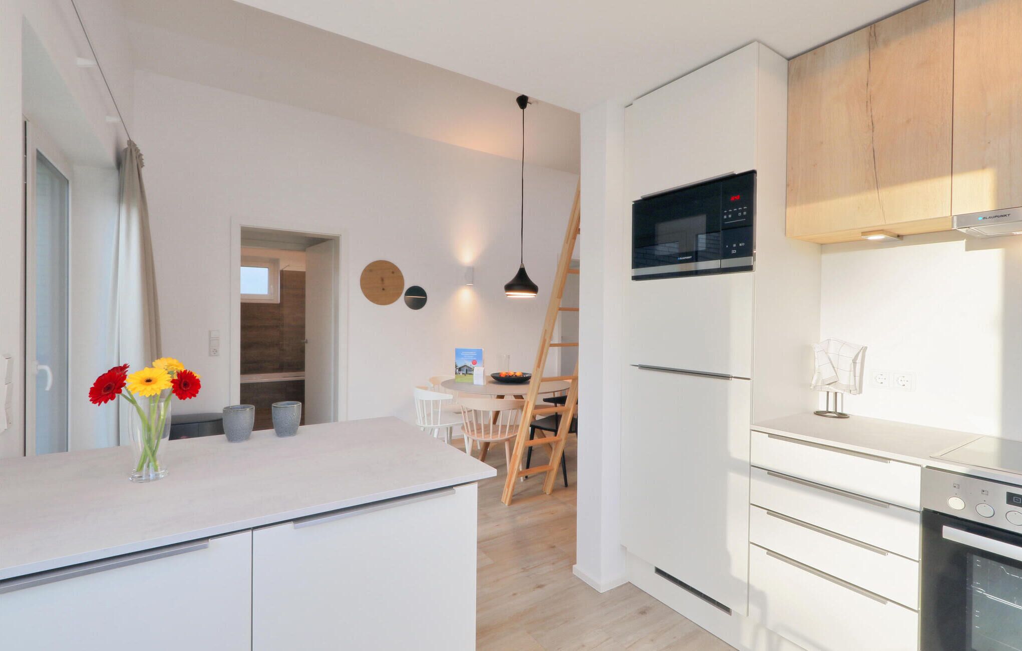 Een moderne keuken met witte kasten, een ingebouwde oven en een magnetron. Er is een eethoek met een houten tafel en stoelen, en een vaas met bloemen op het aanrecht.