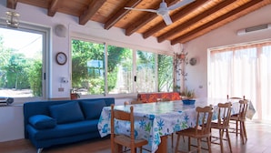 A dining area with a table covered in a floral tablecloth, surrounded by wooden chairs, a blue sofa, and large windows with a view of greenery outside. - Villa Bruffalori (Scicli)