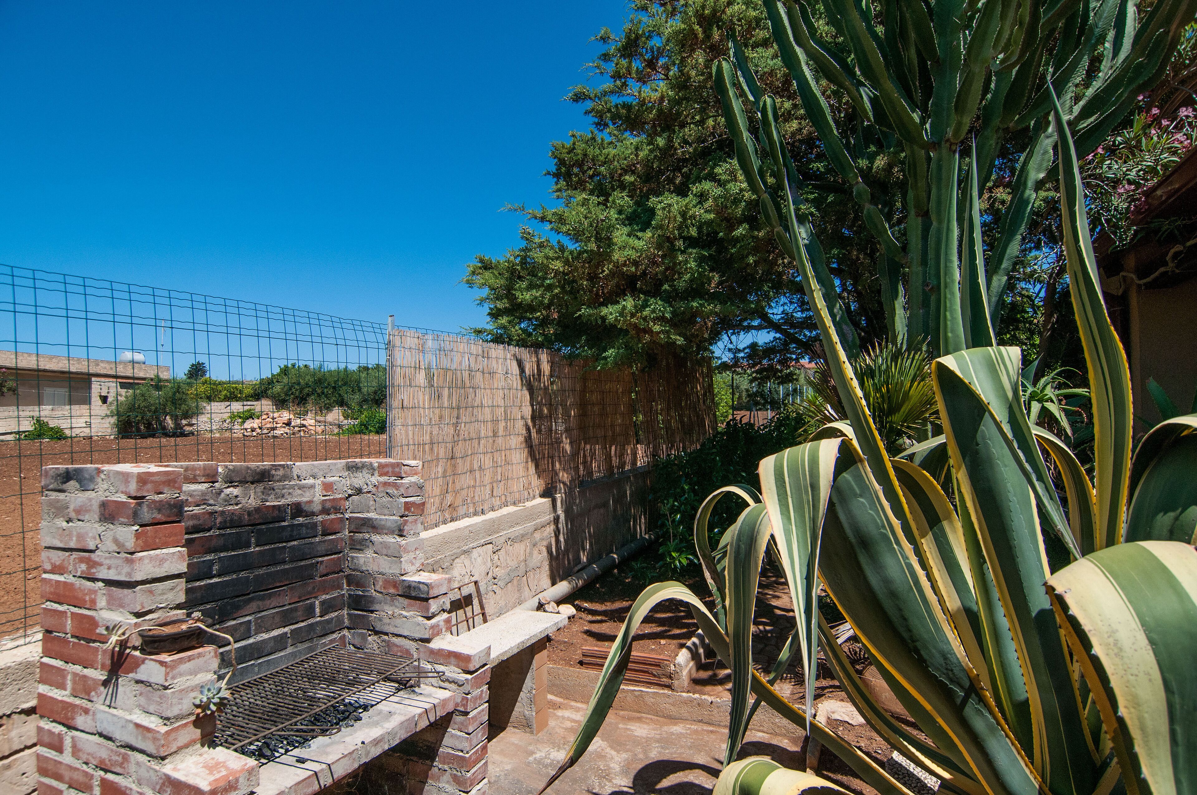 A brick barbecue grill with a metal grate, surrounded by large green plants and a brick wall.