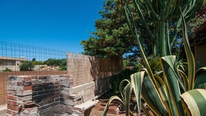 A brick barbecue grill with a metal grate, surrounded by large green plants and a brick wall. - Villa Bruffalori (Scicli)