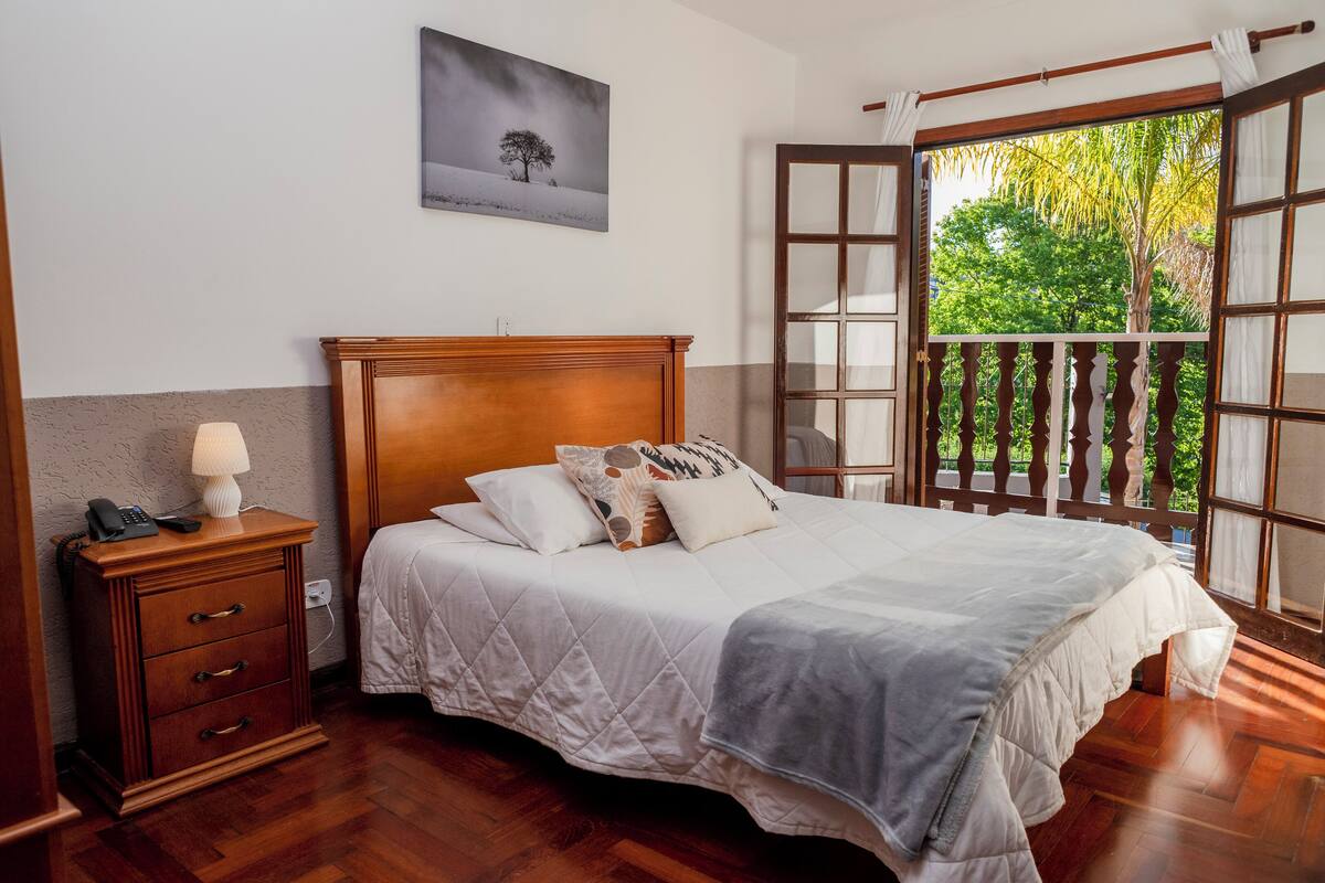 Un dormitorio con una cama de madera, una mesita de noche con lámpara, un teléfono en la pared y una ventana con vistas a zonas verdes.