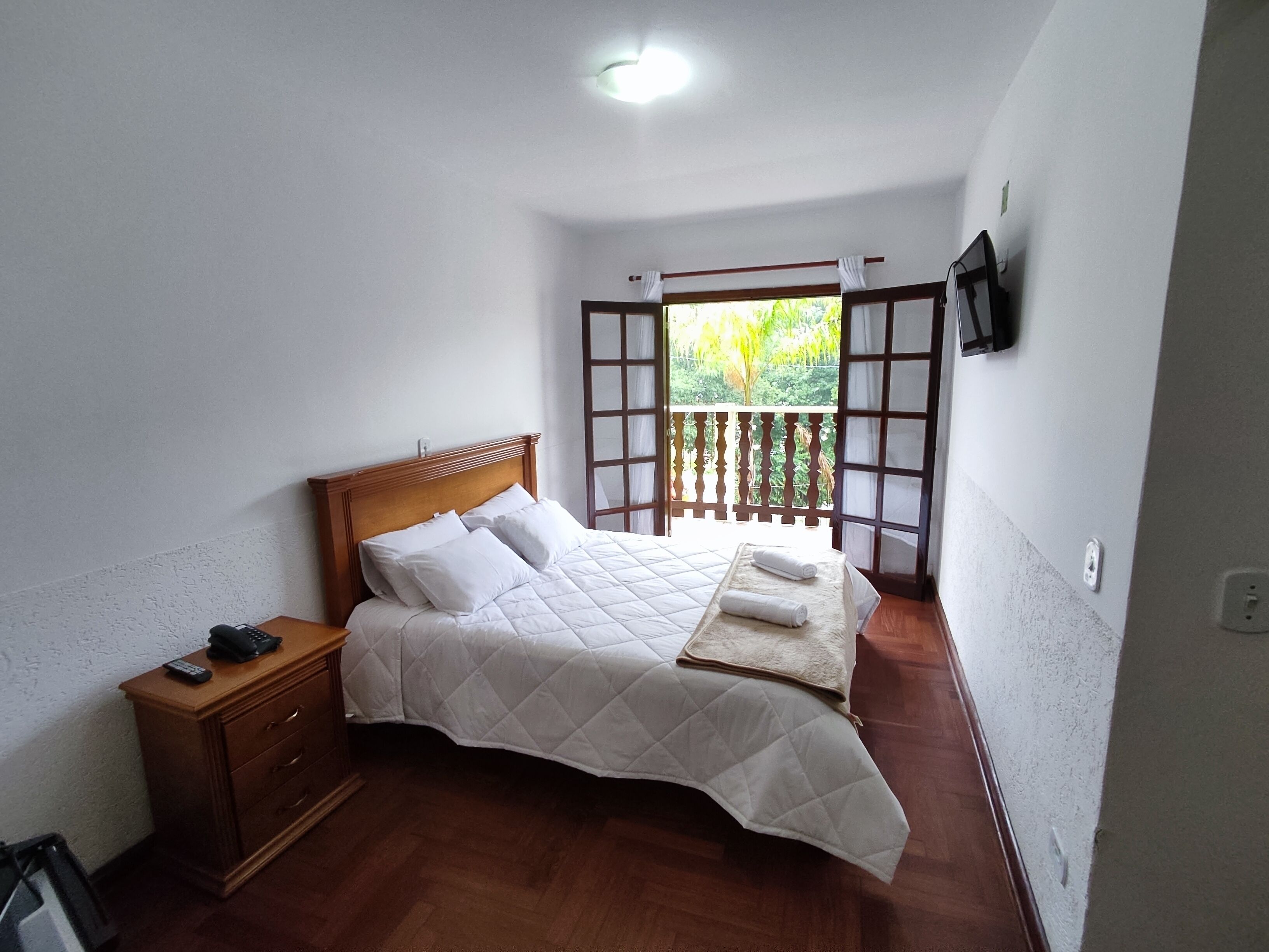 A bedroom with a wooden bed, a nightstand, a TV on the wall, and a balcony with a view of greenery.