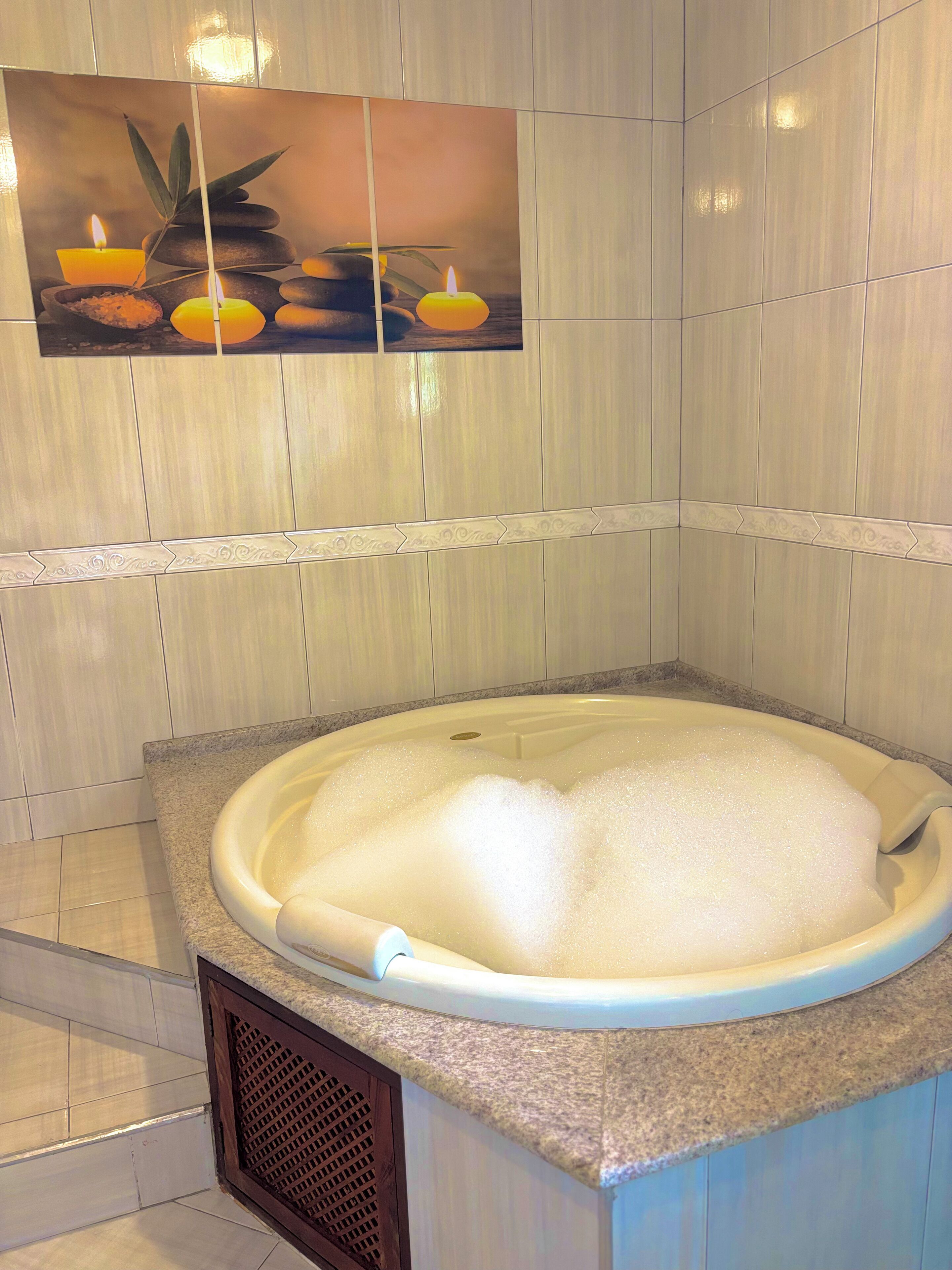 A spa area with a hot tub filled with bubbles, a wall-mounted artwork of candles and stones, and a tiled wall with a decorative border.