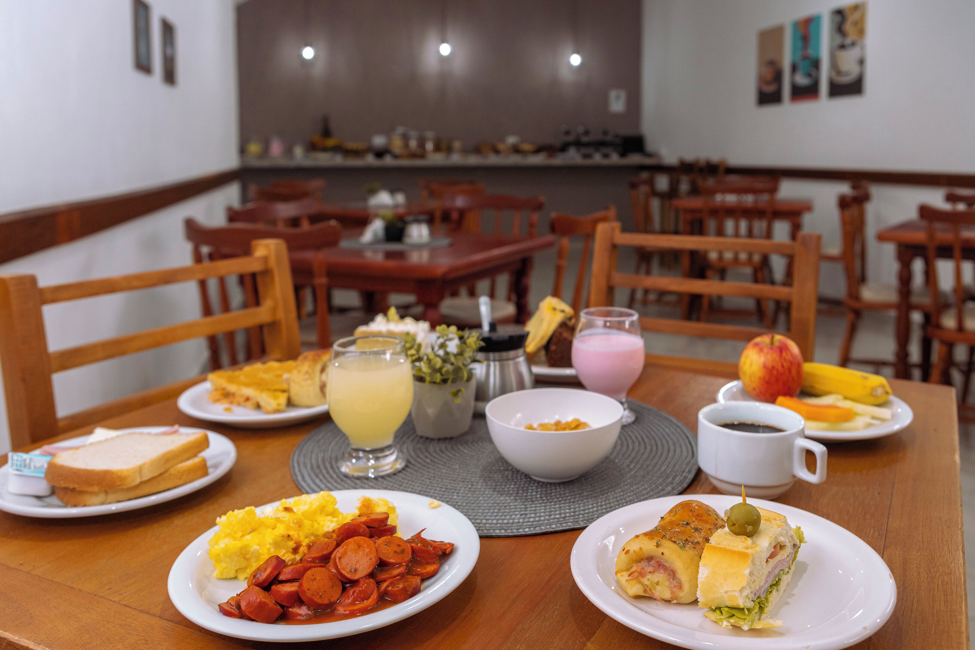 Een ontbijttafel met eten zoals toast, roerei, worst en een belegd broodje. Ook fruit, een koffiekop en een glassap.