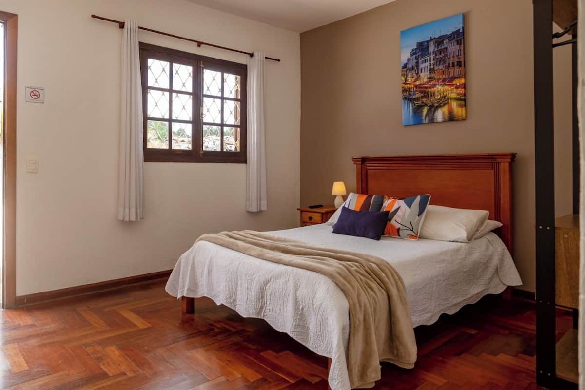 Un dormitorio con una cama de madera, una mesita de noche con una lámpara, una ventana con cortinas y un cuadro de un paisaje urbano en la pared.