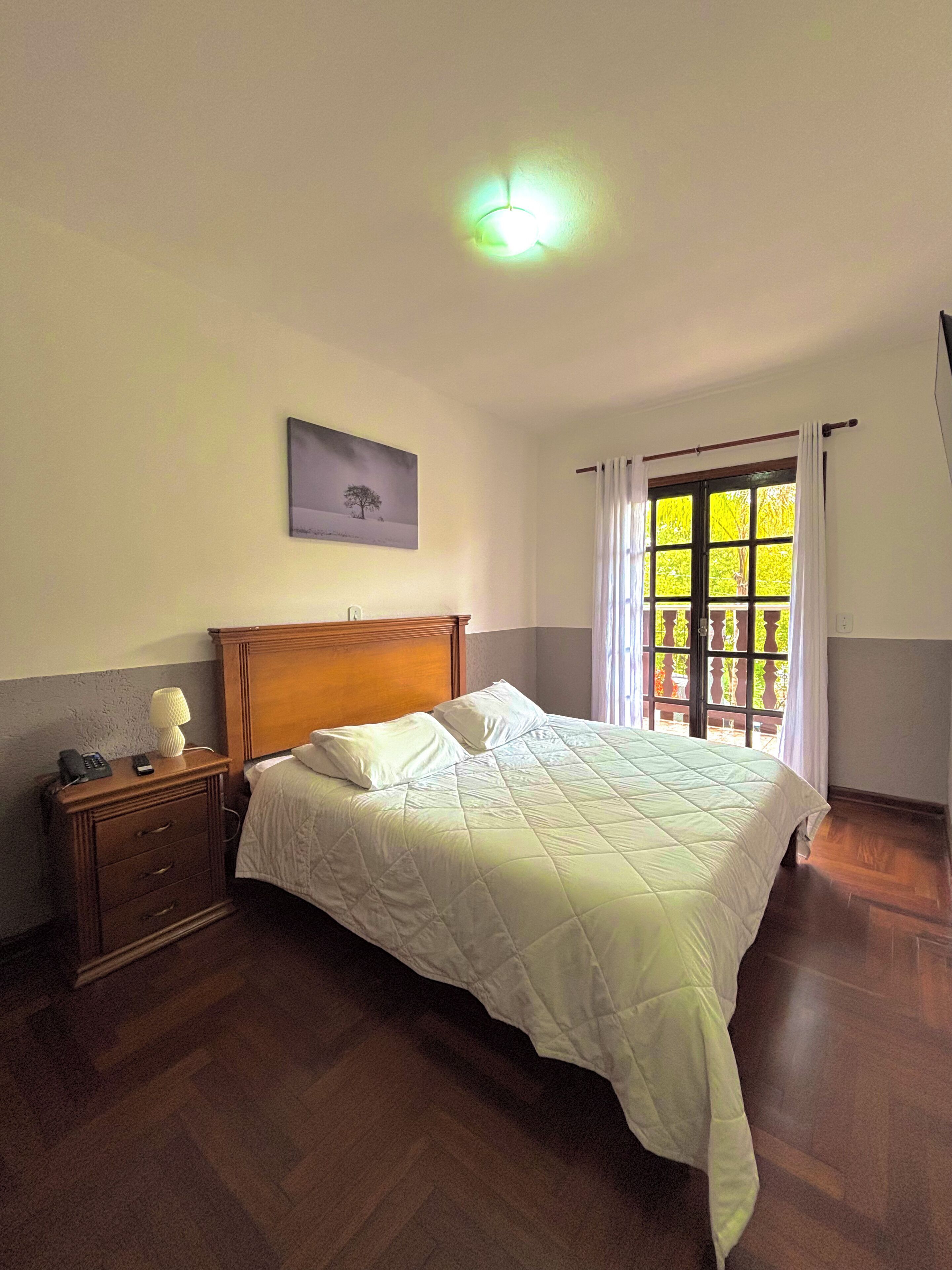 A bedroom with a wooden bed, a nightstand, a painting on the wall, and a window with curtains.