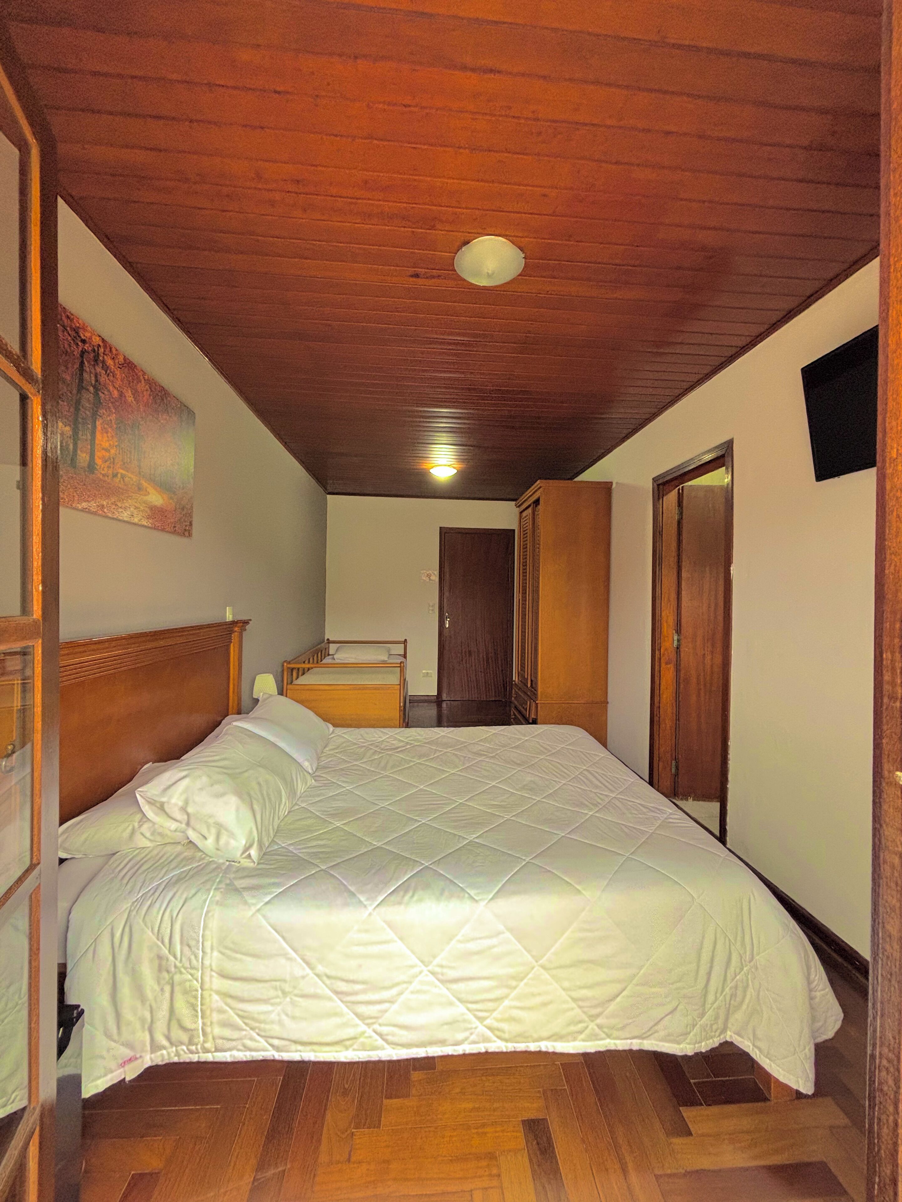 A bedroom with a wooden ceiling, a bed with a quilted white duvet, a painting on the wall, and a door leading to another room.