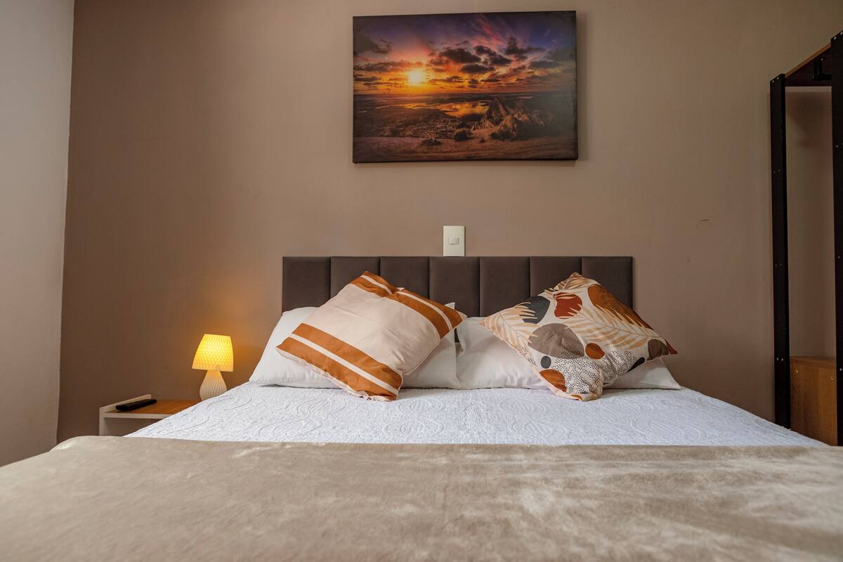 Una cama bien hecha con almohadillas estampadas, una lámpara de noche y un cuadro con un atardecer en la pared.
