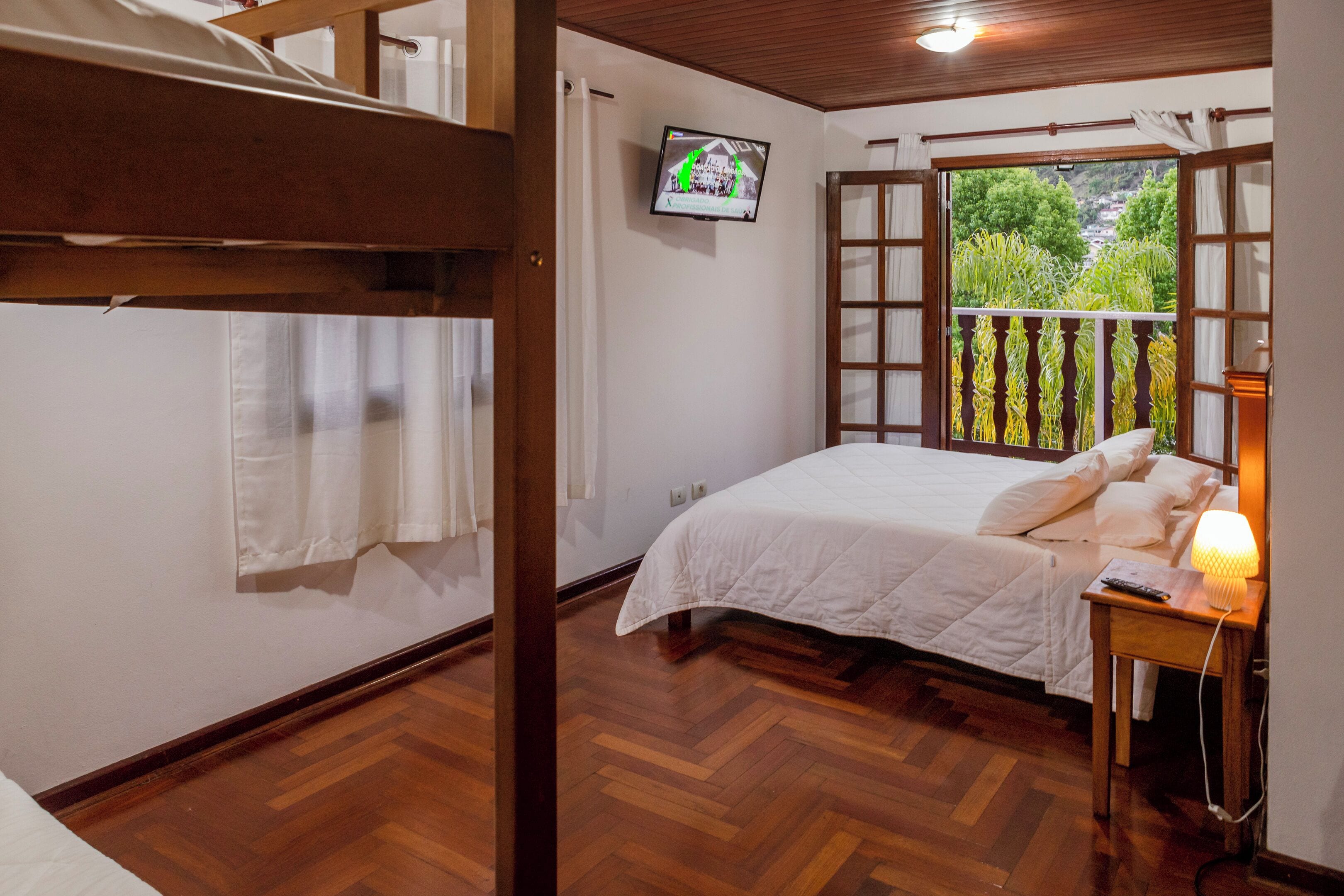 A bedroom with a wooden floor, a bed with white bedding, a TV on the wall, and a balcony with a view of greenery.