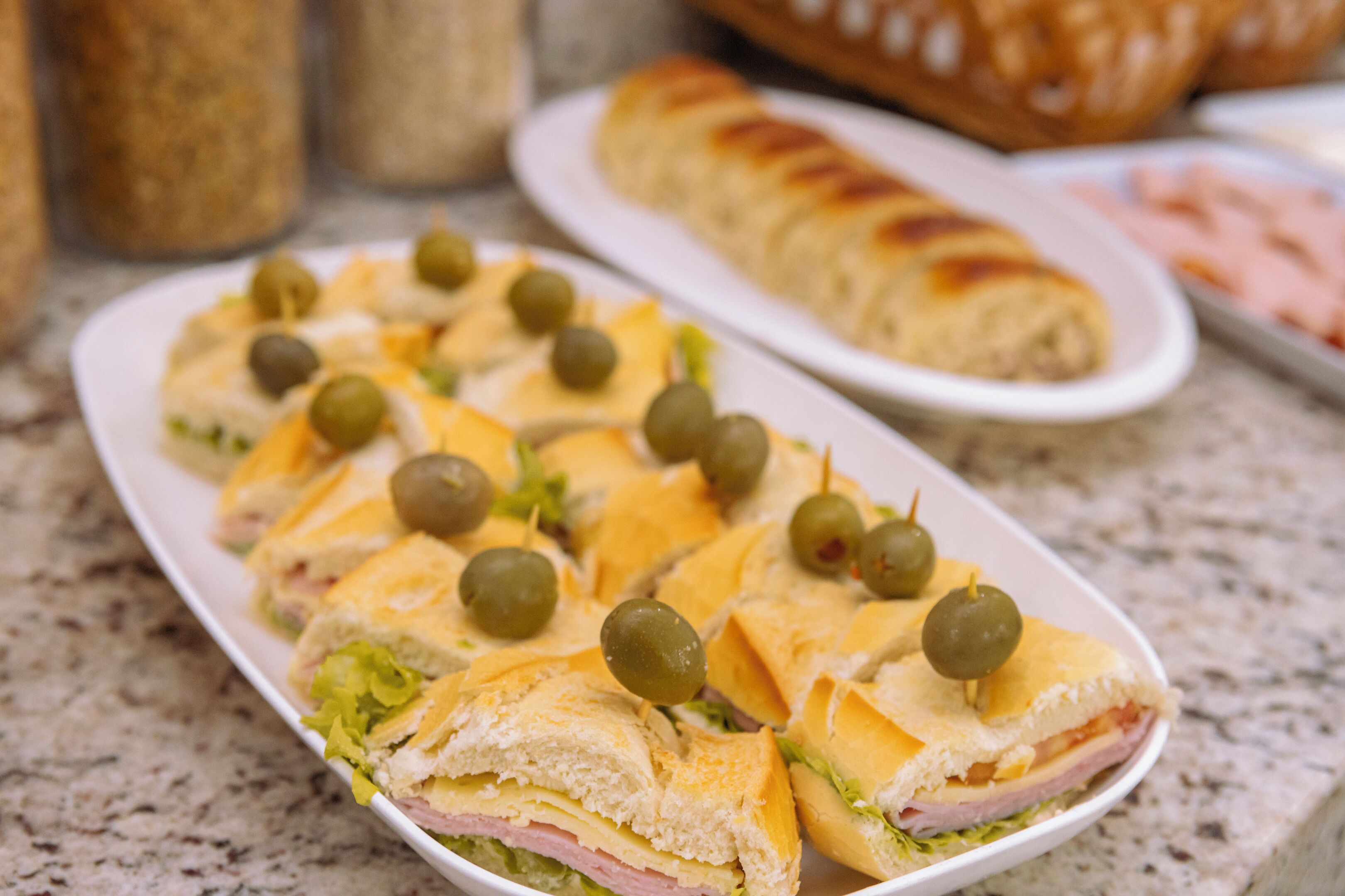 A plate of sandwiches with olives and a loaf of bread in the background.
