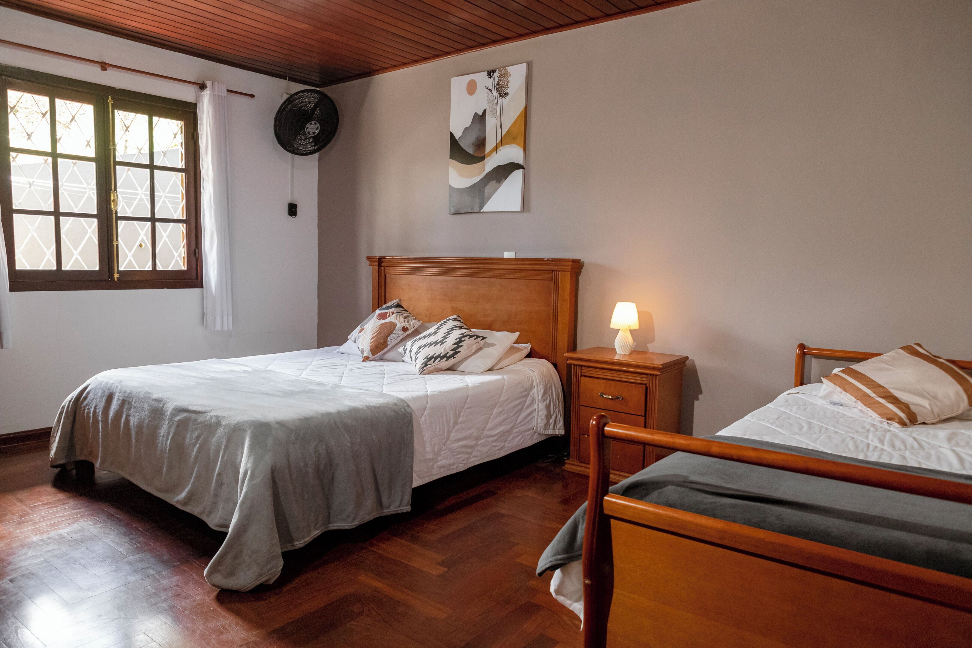 A bedroom with a large bed, a smaller bed, a nightstand with a lamp, a wall-mounted fan, and a window with curtains.