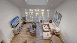 A spacious living room with a large TV, comfortable seating, and a view of the outdoors through large windows.