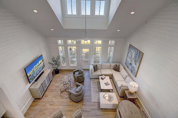 A spacious living room with a large TV, comfortable seating, and a view of the outdoors through large windows.