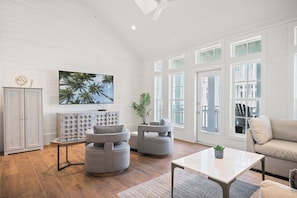 A spacious living room with a high ceiling, a large window with multiple panes, a flat-screen TV mounted on the wall, and a modern grey sofa.