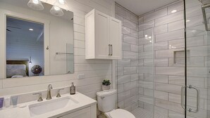 A modern bathroom with a white sink, mirror, and toilet. It features a glass shower enclosure and a small potted plant on the sink.