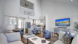 A spacious living area with a high ceiling, a balcony, a kitchen, and a television displaying a pool view.