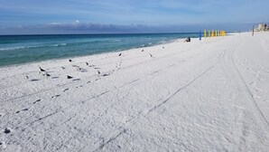 Ein weiter Sandstrand mit Fußspuren, die zum Wasser führen, eine Person in der Nähe von gelben Sonnenschirmen und ein klarer, blauer Himmel.