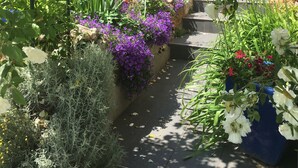 Un jardín con diversas flores y plantas, un área de estar techada al aire libre, y un edificio con contraventanas azules al fondo.