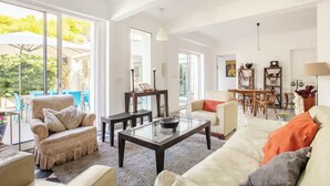 Amplia sala de estar con una mesa de centro de vidrio, sofá color beige y una mesa de comedor con sillas. Amplias ventanas ofrecen luz natural y vistas al exterior.