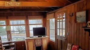 Un accogliente interno di una baita con pareti e pavimento in legno, un divano con cuscini decorati, una scrivania con un televisore e un cartello appeso al muro.