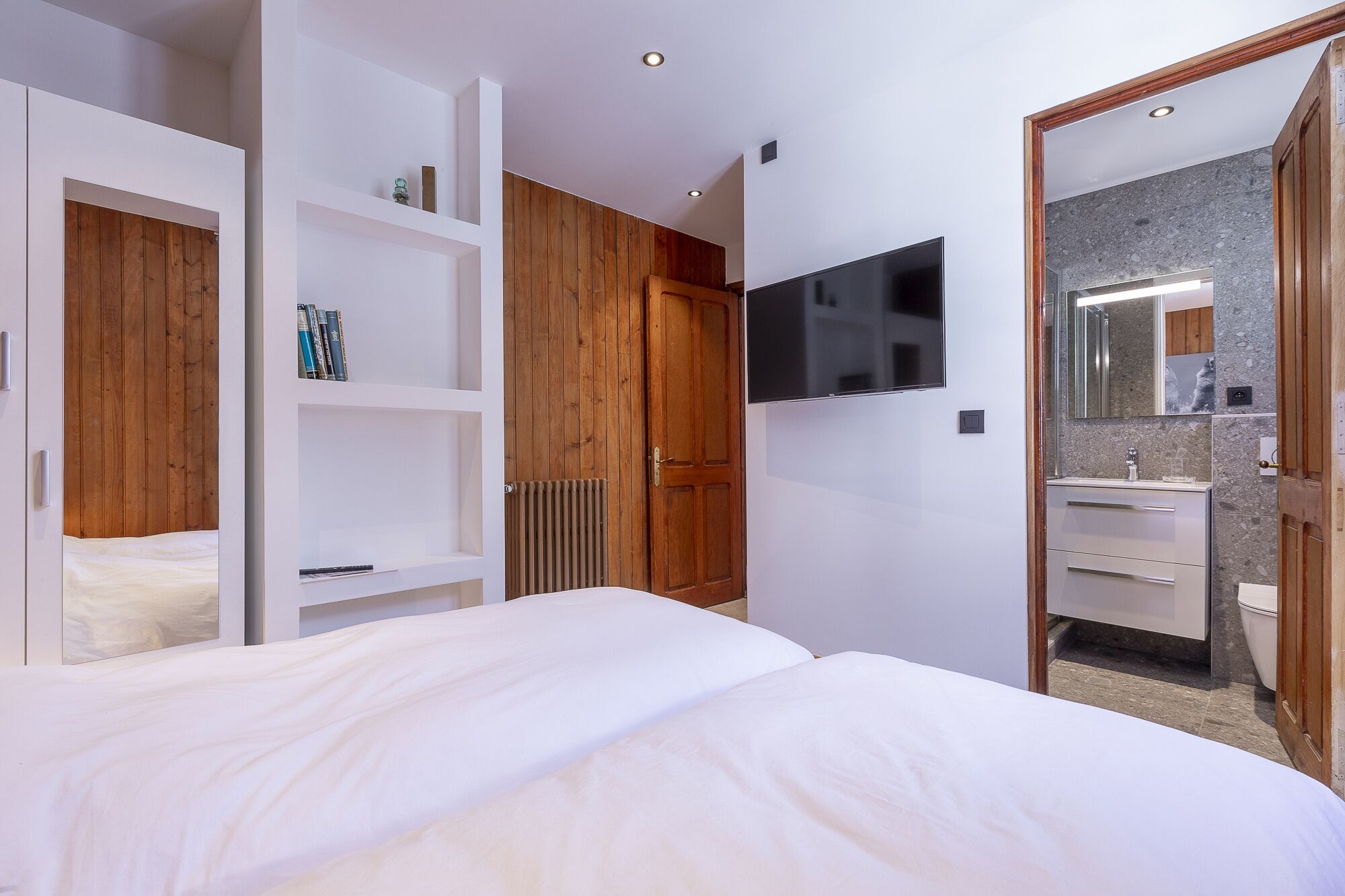 Un dormitorio moderno con una cama grande, estanterías integradas, un televisor y un baño con espejo y lavamanos.