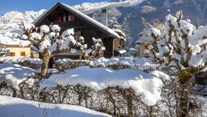 A wooden chalet with a sloped roof, surrounded by snow-covered trees and mountains in the background.