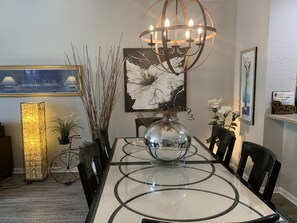 Een eetgedeelte met een tafel met glazen blad, zwarte stoelen, een kroonluchter, een groot abstract schilderij en een vaas met witte bloemen.