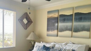 A bedroom with a bed, a ceiling fan, a window with blinds, a lamp, and three landscape paintings on the wall. - Short/Long Term Fully Furnished Rental (Carlsbad)