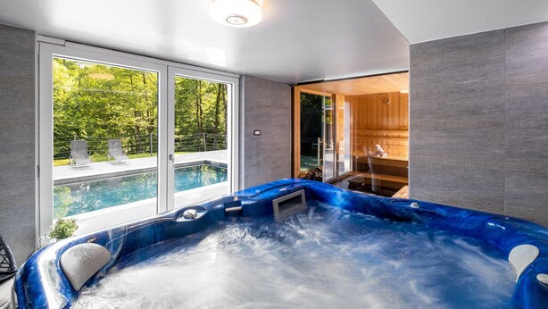 A modern indoor swimming pool with a hot tub, large windows, and a view of a forested area.