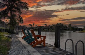 A waterfront deck with chairs and a dock extending over calm water, palm trees, and a sunset sky.
