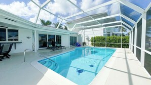 En indendørs swimmingpool med krystalklart blåt vand, omgivet af glas, og en terrasse med stole og et bord.