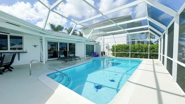 An indoor swimming pool with clear blue water, surrounded by a glass enclosure, and a patio area with chairs and a table.