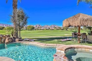 A pool area with clear water, a thatched umbrella, and a view of a golf course with mountains in the background.