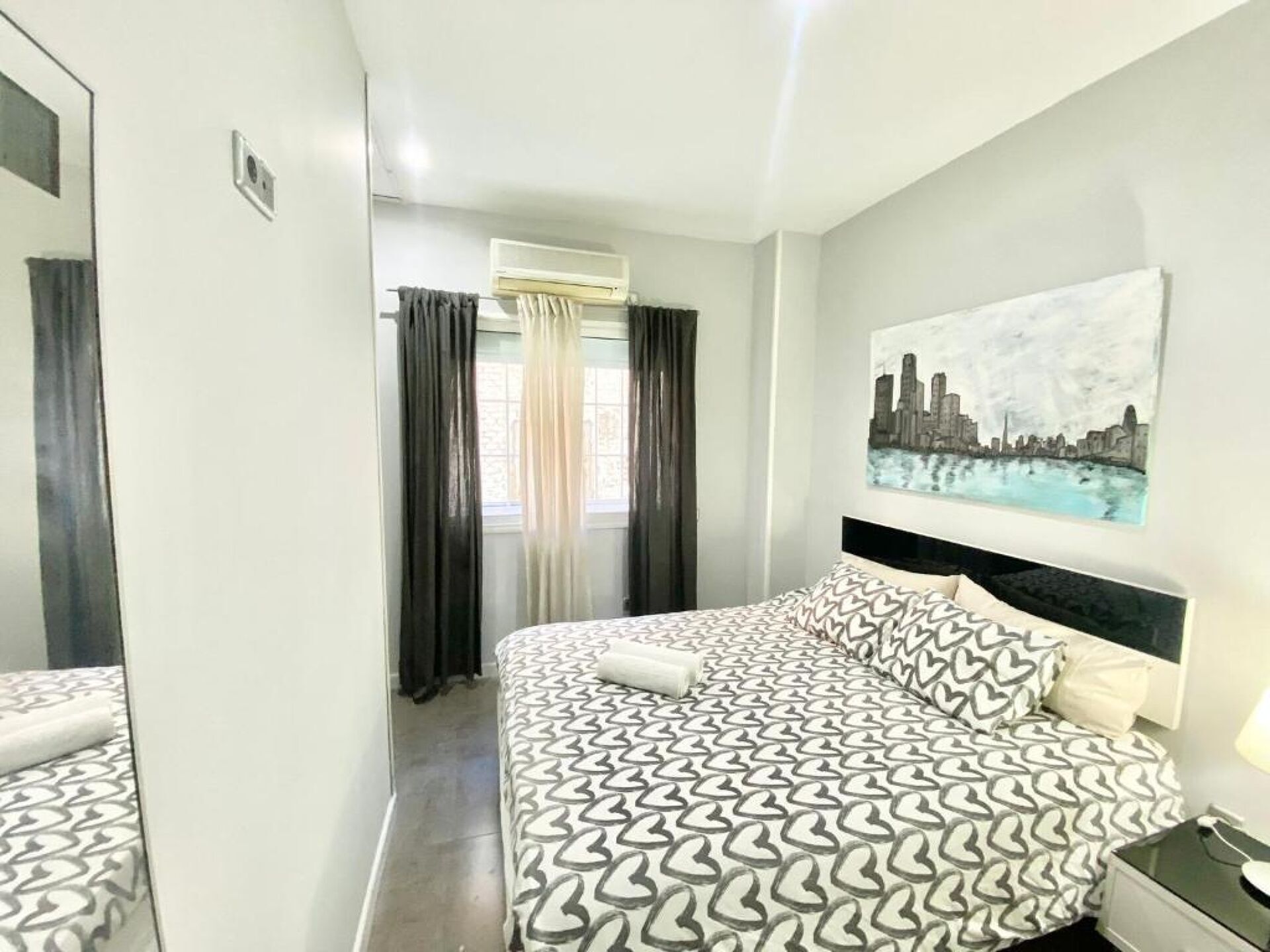A hotel room with a bed, a nightstand, a painting on the wall, and a window with curtains.